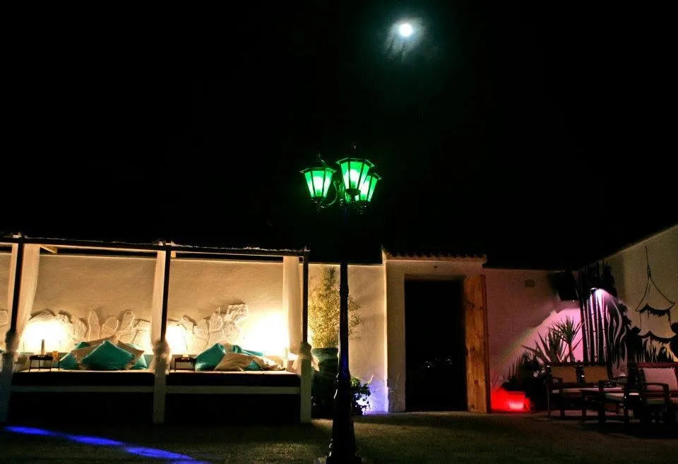 Terraza de noche con una farola verde, camas balinesas iluminadas con cojines turquesa y la luna al fondo